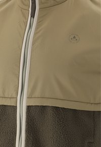 Jacka med dragkedja, där den övre delen är ljusbrun och den nedre delen är mörkbrun fleece. Den har en rund logo på övre vänstra sidan.