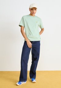 Maglietta di cotone verde chiaro con un piccolo logo, abbinata a jeans scuri a gamba larga e scarpe da ginnastica blu. Un cappello beige completa l'outfit.