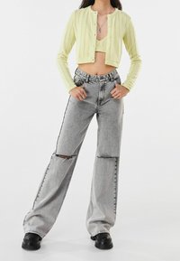 Kobieta ubrana w krótki żółty sweterek i braletkę, szare, wysokowziaste, szerokie jeansy z rozdarciami oraz czarne masywne buty.