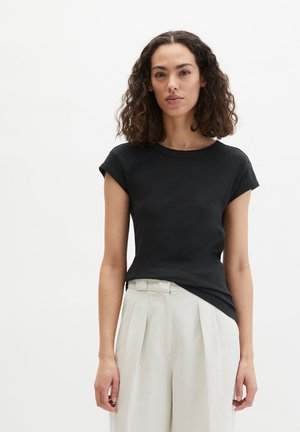 Kobieta o kręconych włosach, ubrana w dopasowaną czarną koszulkę z krótkim rękawem oraz kremowe plisowane spodnie z wysokim stanem na jednolitym białym tle.