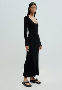 Schwarzes, geripptes Langarmkleid mit tiefem Rundhalsausschnitt und ausgestelltem Saum. Getragen mit schwarzen Plattform-Flip-Flops, die eine elegante Silhouette zur Schau stellen.