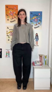 Ženska v sivi majici z dolgimi rokavi in črnih hlačah stoji pred steno s pisanim risanim plakatom in belo predalno omarico s knjigami.