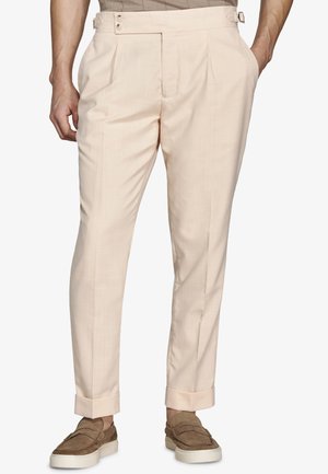 Pantaloni beige su misura con linguette regolabili laterali, orli risvoltati, indossati con mocassini in suede marrone su una persona in piedi.