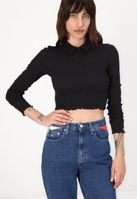 Svart avskåret langermet polo med knappelukking og bølget kant, parret med høyhående blå denimjeans med frontlommer.