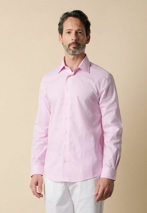 Mann mit kurzen dunklen Haaren und Bart, trägt ein hellrosa gestreiftes Hemd mit Knöpfen und weiße Hose vor beigefarbenem Hintergrund.