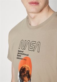 Beige bomulds t-shirt med sort NASA-logo og tekst, samt en orange og grå grafik af månen og en kredsende satellit.