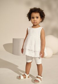 White layered dress with perforated detailing, ribbed white shorts, and white sandals with ankle strap. Smooth fabric and textured accents.