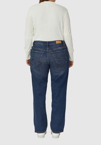 Jean en denim bleu foncé à coupe décontractée, avec deux poches arrière et des accents de couture claire. Porté avec un haut blanc à manches longues.