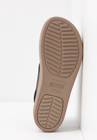 Semelle d'une chaussure Crocs bronzée avec motifs de la bande de roulement, positionnée à la verticale contre un mur blanc d’angle, montrant le nom de la marque et les détails de la taille.