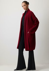 Nő hosszú bordó kardigánban, fekete garbóval és bőszárú nadrággal, oldalról áll, az egyik kezét a zsebében tartva, sima háttér előtt.