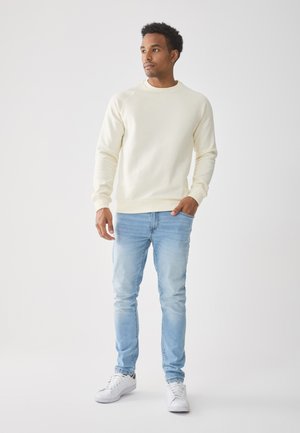 Mann steht mit einer Hand in der Tasche, trägt einen cremfarbenen Pullover, hellblaue Jeans und weiße Sneaker vor einem schlichten weißen Hintergrund.