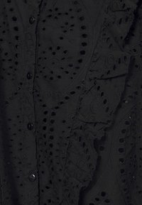 Tissu noir avec une broderie ajourée complexe en motifs tourbillonnants, avec de petits boutons ronds et des détails délicatement froncés.