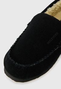 Černé semišové pantofle s měkkou kožíškovou podšívkou. Obsahují prošívání kolem okraje a vyražené logo na boku. Ploché podešev pro pohodlí.