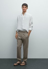 Jonge man in een wit overhemd met knopen, beige broek en zwarte teenslippers, staand met één hand in zijn zak tegen een effen achtergrond.