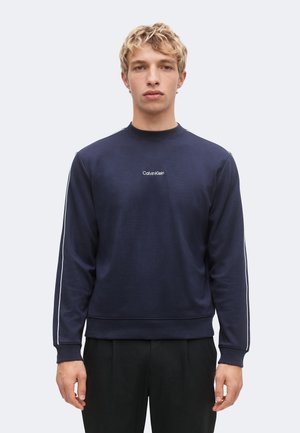 Młody mężczyzna z krótkimi blond włosami, ubrany w granatową bluzę Calvin Klein z białymi paskami po bokach, stoi na jasnym, jednolitym tle.