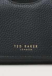 Crna teksturirana kožna torbica sa zlatnim natpisom "TED BAKER LONDON", glatkim završetkom i zaobljenim gornjim rubom.