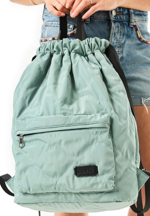 Hellgrüner gesteppter Rucksack mit Kordelzug und vorderer Reißverschlusstasche, von Händen über Jeansshorts gehalten.