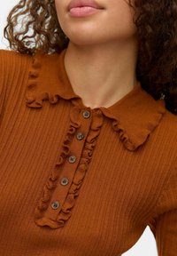 Gros plan sur une personne portant un pull en maille côtelée couleur rouille avec un col volé et une patte de boutonnage, cheveux frisés partiellement visibles.