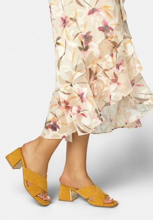 Pies con sandalias mule de tacón bloque en color mostaza combinadas con una falda floral transparente de longitud media en tonos suaves de beige, rosa y marrón.