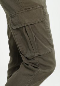 Gros plan d'un pantalon cargo vert olive avec une grande poche latérale à rabat sur la cuisse et des détails de couture visibles.