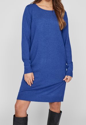 Mujer con un vestido suéter azul de manga larga y largo hasta la rodilla, combinado con botas negras hasta la rodilla, sobre un fondo gris liso.