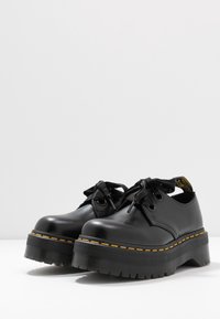 Scarpe in pelle nera con punta arrotondata, suola piatta spessa e cuciture gialle a contrasto. Presentano lacci piatti neri e un tab sul tallone nero.