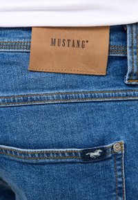 Blaue Jeans aus Denim verfügen über ein braunes Lederpatch mit der Aufschrift "MUSTANG" am Bund. Enthält gelbe Nähte und ein kleines Logo auf der Gesäßtasche.
