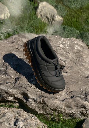 Sort isoleret slip-on sko med tyk brun sål med mønster, hvilende på en solbeskinnet klippe med græs i baggrunden.