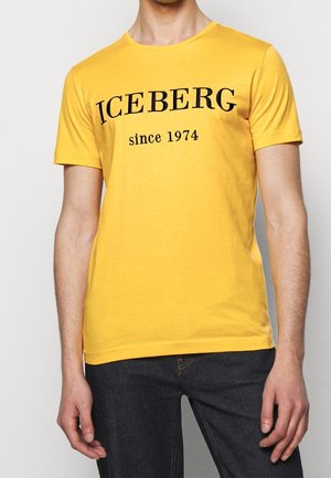 Homme portant un t-shirt jaune avec le texte noir "ICEBERG since 1974" associé à un jean bleu foncé, debout devant un fond uni.