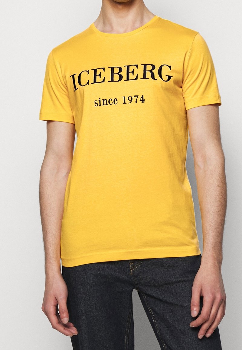 Homme portant un t-shirt jaune avec le texte noir "ICEBERG since 1974" associé à un jean bleu foncé, debout devant un fond uni.