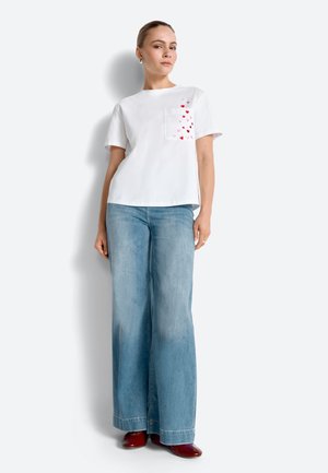 Žena stojící, oblečená v bílé krátkosleevé košili s malými červenými srdíčky na kapse, širokých modrých džínách a červených botách na bílém pozadí.