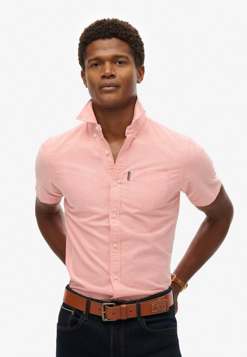 Camicia a maniche corte rosa chiaro con colletto button-down, taschino sul petto e una sottile textura. Abbinata a una cintura marrone e jeans scuri.