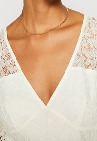 Vit spetsblus med djup V-halsringning och blommönster. Korta ärmar med spetsdetaljer, kompletterad med ett tunt guldfärgat halsband.