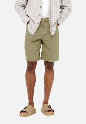 Homme portant un short olive mi-genou, des sandales beiges à bouts ouverts, une chemise blanche et une chemise beige à carreaux boutonnée, les mains dans les poches.