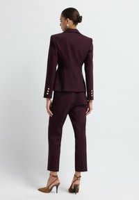 Femme avec les cheveux en chignon portant un tailleur ajusté de couleur violet foncé avec des boutons dorés et des chaussures à talons hauts marron, se tenant devant un fond clair uni.