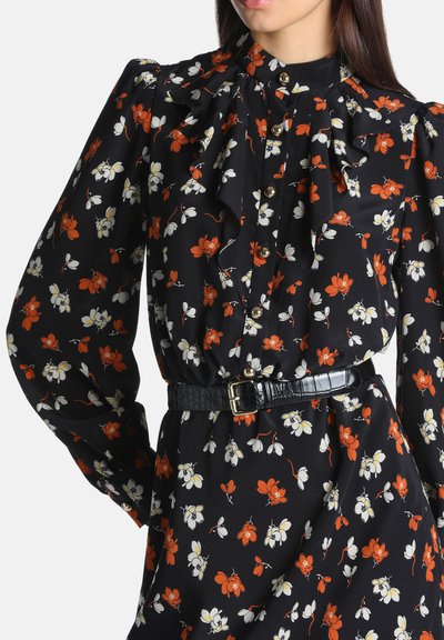 Femme portant une robe noire à fleurs rouges et blanches, boutons dorés, devant froncé et ceinture noire avec boucle dorée.