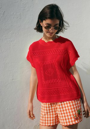 Rövid sötét hajú nő, kerek napszemüveget visel, piros texturált kötött felsőt és narancssárga kockás rövidnadrágot, hullámos szegéllyel és hímzéssel.