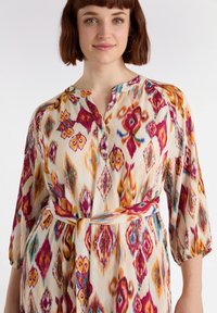 Monivärinen, kuvioitu mekko, jossa on kaulus, napitus ja sidottava vyö. Sisältää kirkkaita sävyjä vaaleanpunaisesta, oranssista ja tealista kermansävyisellä taustalla.
