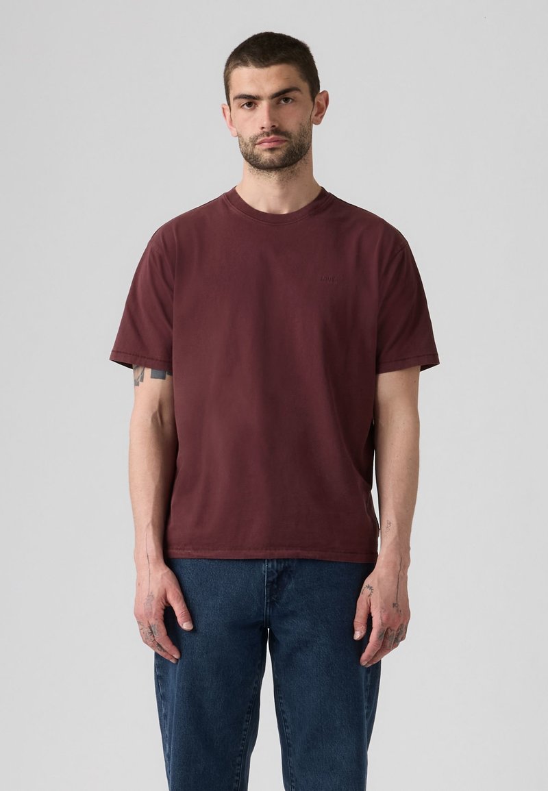 Uomo con capelli corti scuri e barba che indossa una maglietta bordeaux tinta unita e jeans blu scuro, in piedi davanti a uno sfondo grigio chiaro.