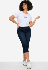 Maglietta bianca con la scritta "Capri", jeans cropped blu scuro e sneaker bianche. Il modello ha capelli ricci e occhiali oversize.
