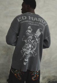 Maglione grigio a coste con design grafico di teschio e tibie sul retro. Presenta un collo a giro e spalle scese.