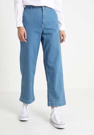 Kobieta nosząca niebieskie jeansy z wysokim stanem i szerokimi nogawkami, w połączeniu z białymi, wysokimi sneakersami i białą koszulą z długim rękawem, stojąca na białej podłodze.