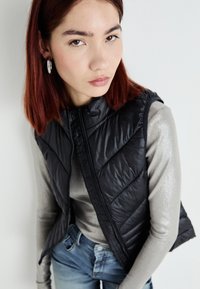 Black padded vest with a zip closure, featuring a diamond quilted pattern, worn over a silver long-sleeve top and blue jeans.