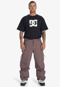 Pantalons de neige gris avec des zippers sur les côtés, associés à un t-shirt noir avec le logo DC. La tenue comprend des baskets blanches et noires avec des accents jaunes.