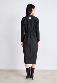 Schwarzes langärmliges Kleid mit tailliertem Oberteil, plissierter Textur und Midi-Länge. Kombiniert mit kniehohen schwarzen Absatzstiefeln.
