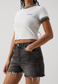 Biały krótki t-shirt z zielonym i czarnym wykończeniem, z szarym logo "LEVI’S", zestawiony z postrzępionymi czarnymi szortami jeansowymi.