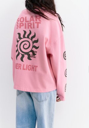 Persoană purtând o cămașă roz cu mâneci lungi, având un spiralat negru reprezentând soarele și textul „SOLAR SPIRIT”, și blugi bleu deschis, văzută din spate.