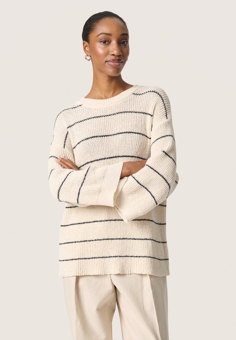 Cremefarvet sweater med marineblå horisontale striber, ribbet tekstur, brede ærmer og rund udskæring, parret med lyse bukser.