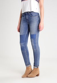 Jeans skinny de denim azul con diseño de parches. Presenta diferentes tonos de azul y detalles desgastados. Combinados con botas de tobillo marrón claro.