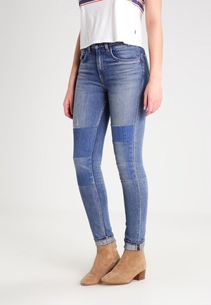 Jeans skinny de denim azul con diseño de parches. Presenta diferentes tonos de azul y detalles desgastados. Combinados con botas de tobillo marrón claro.